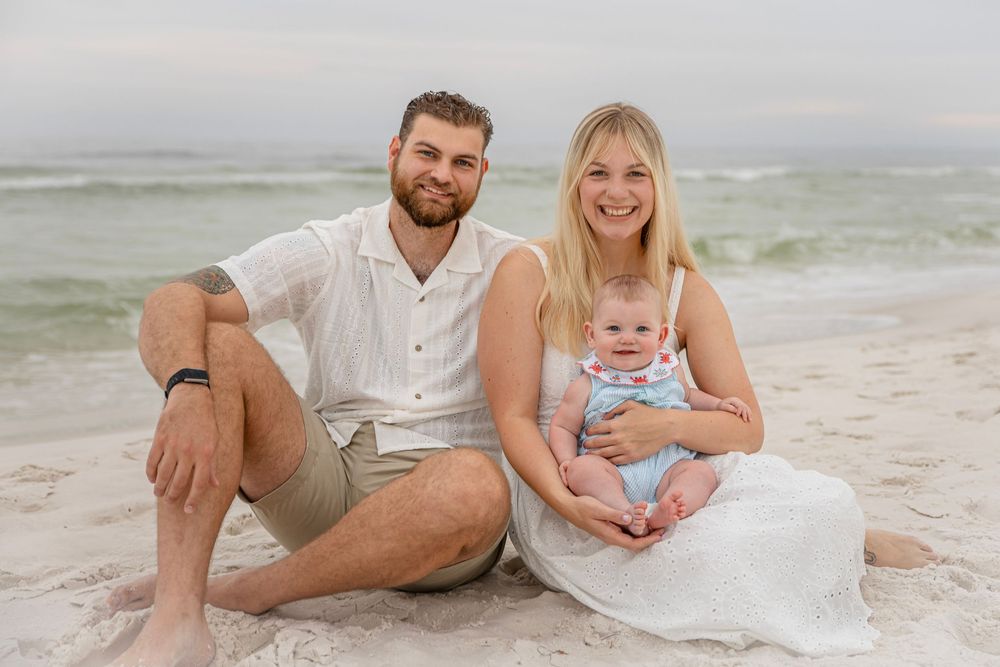 Young family with baby poses on sandy beach, smiling couple in white outfits holding infant by ocean waves.