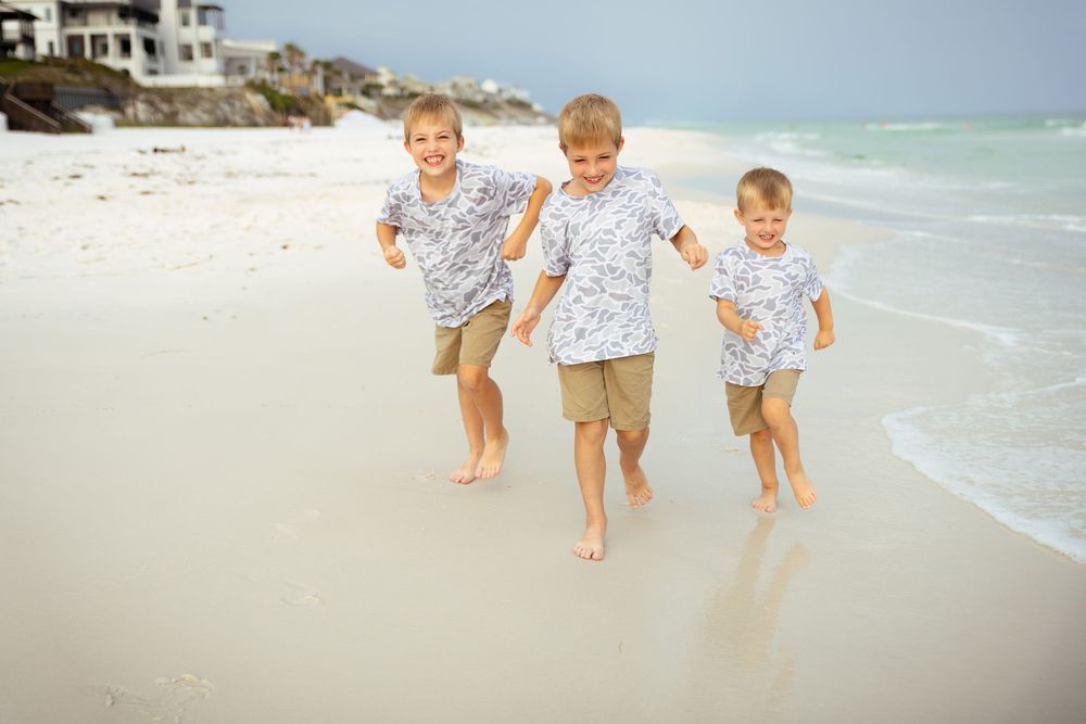 Three boys in matching floral shirts and khaki shorts running joyfully on a white sandy beach near ocean waves.