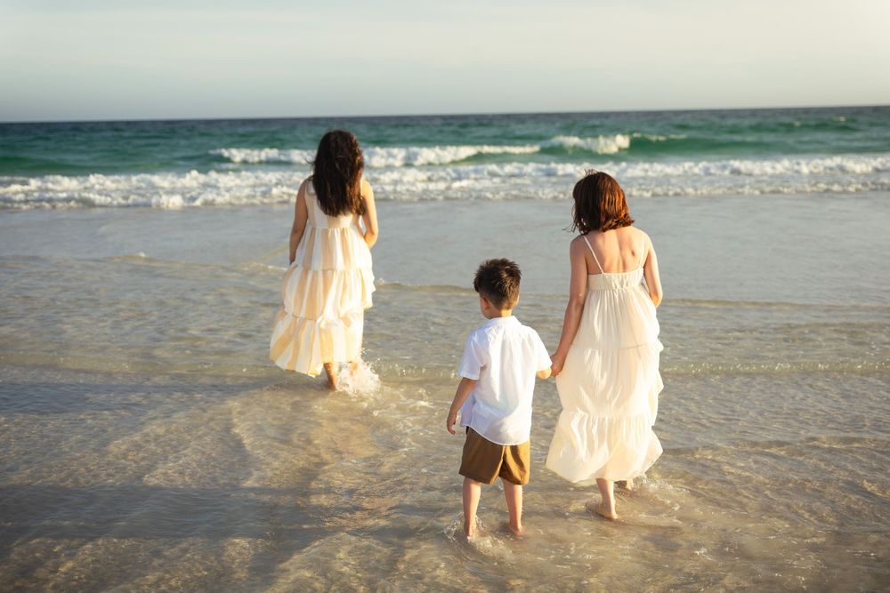 Two women in white dresses and a young boy walk hand-in-hand along a sunny beach with turquoise waves.