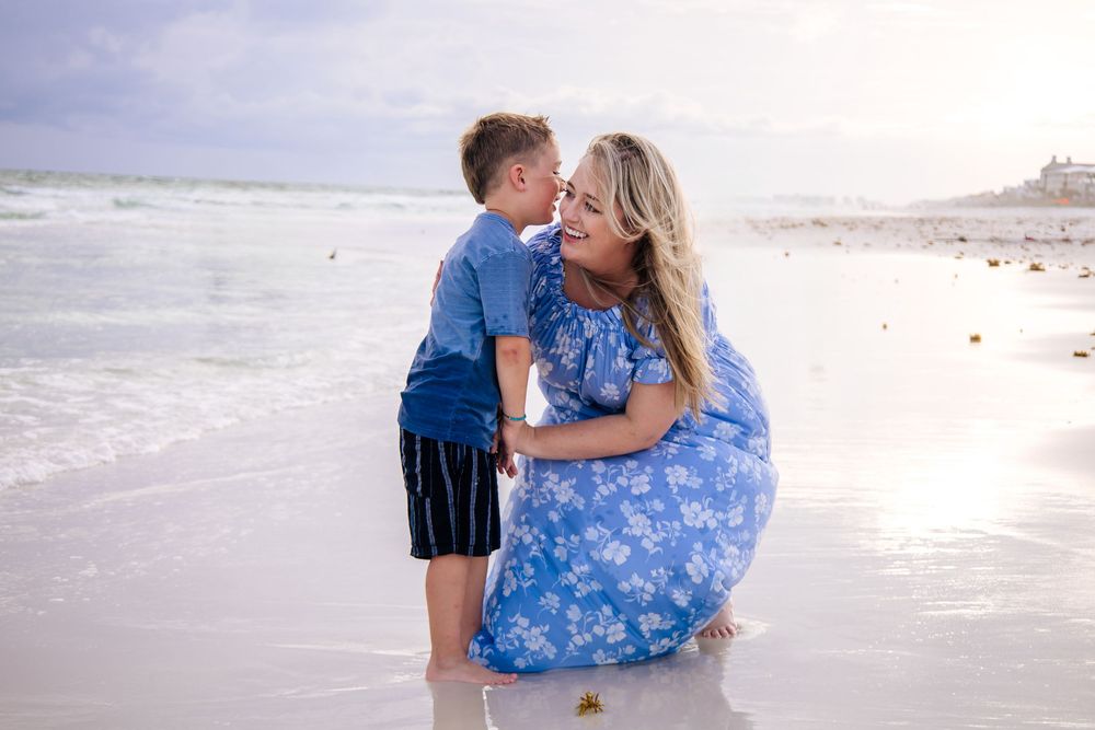 A mother in a blue floral dress kneels on a sandy beach, sharing a tender moment with her young son by the ocean.