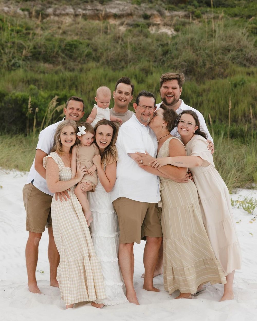 Large family group photo on white sand beach with green dunes backdrop, wearing neutral tones.