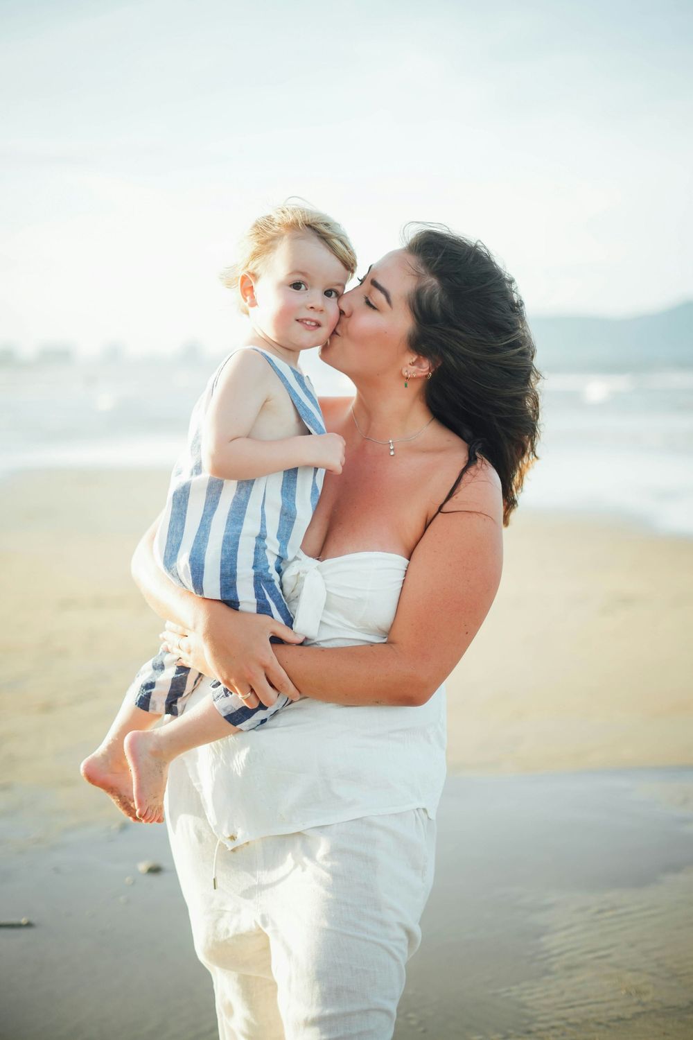 Mother kissing smiling toddler in striped outfit on sunny beach shoreline.