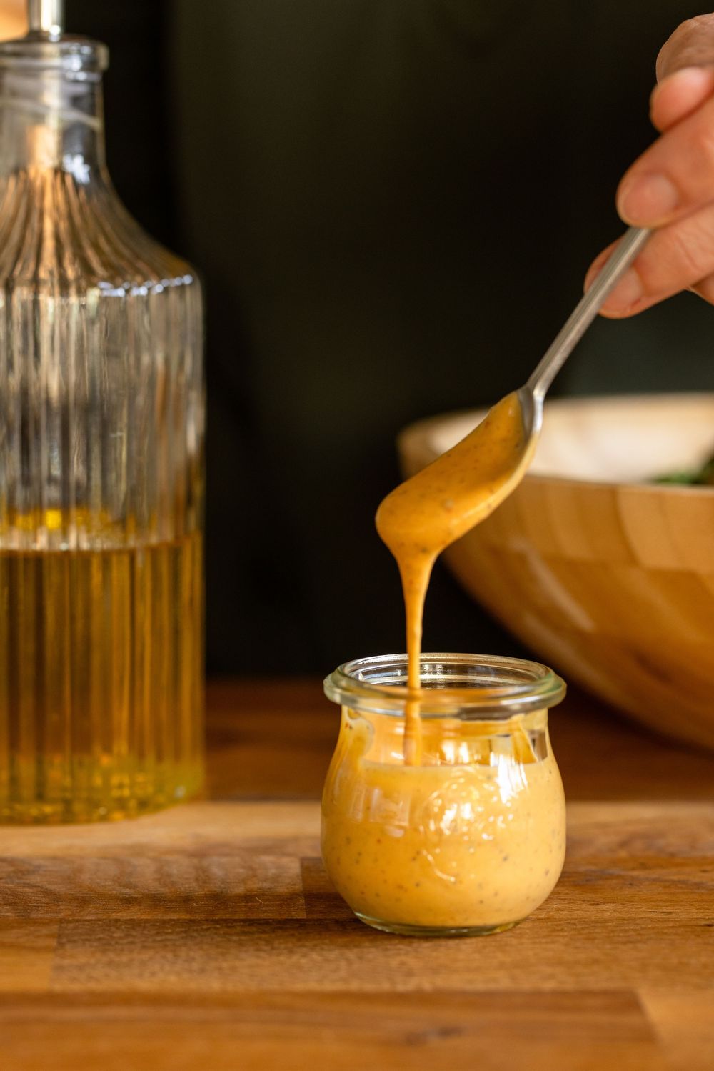 A spoon drizzling homemade mustard dressing into a small glass jar on a wooden surface beside an oil bottle.