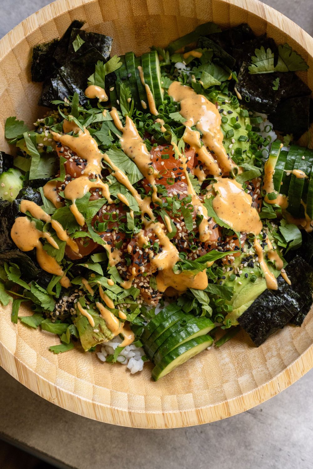 Poke bowl in bamboo bowl with tuna, greens, avocado, nori, sesame seeds, and spicy mayo drizzle.