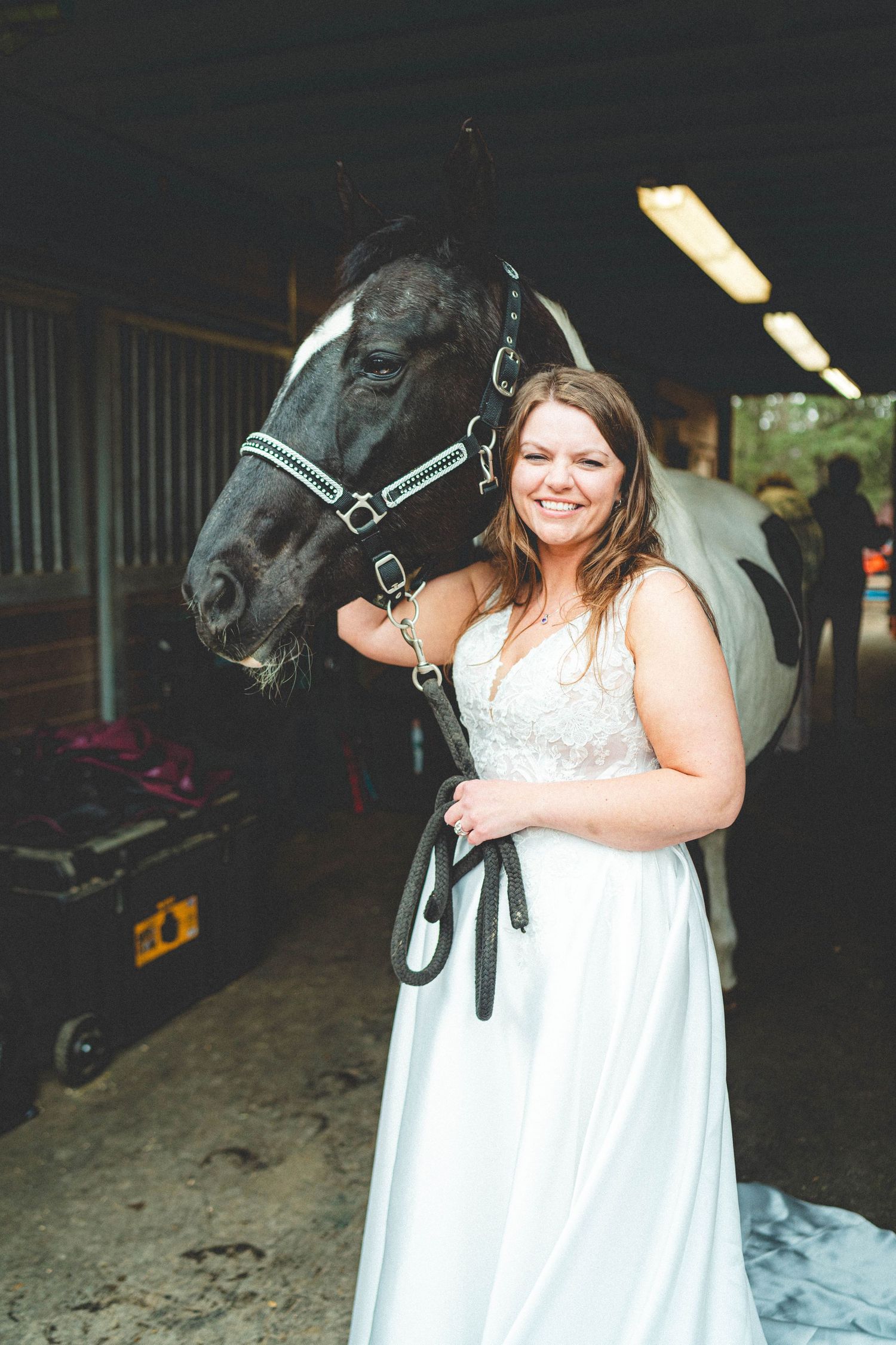 Smiling bride in white gown posing with a black and white horse inside a stable.