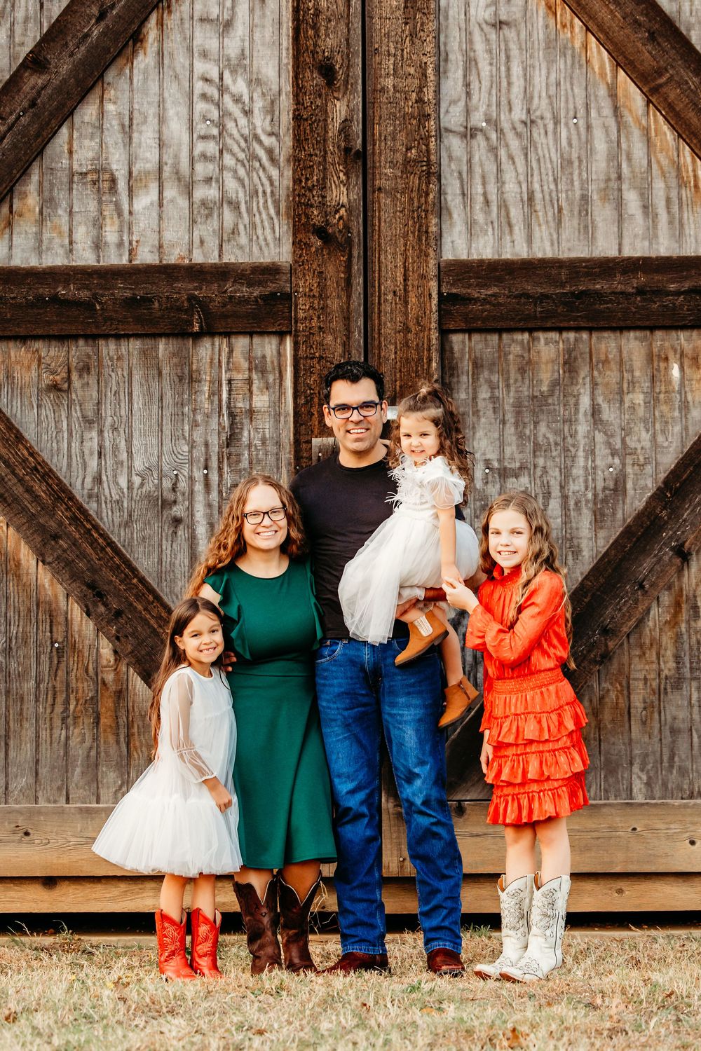 Family of five posing in front of rustic barn doors, wearing coordinated outfits with cowboy boots for a country photo session.
