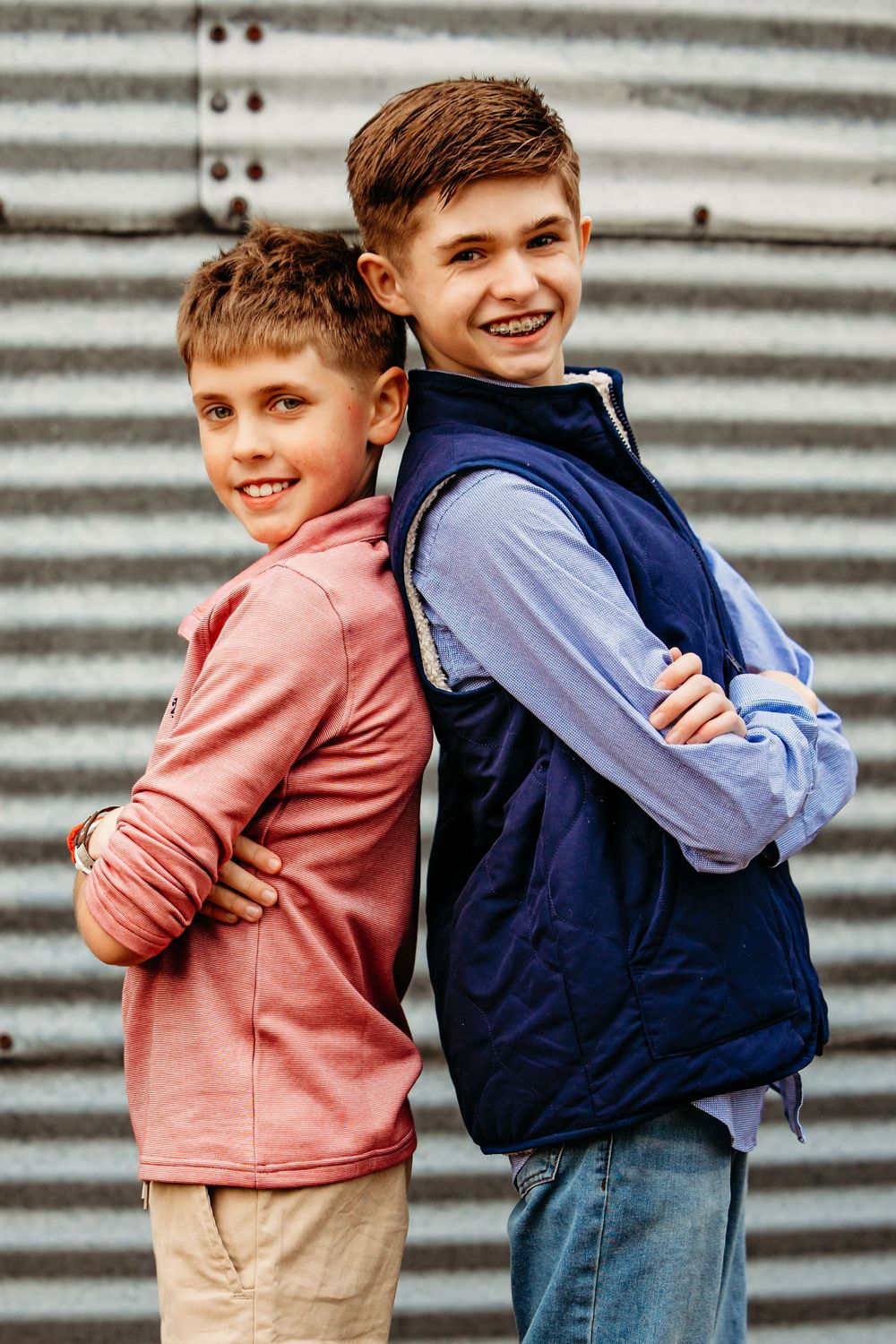 Two smiling boys standing back-to-back with arms crossed in front of a corrugated metal wall.