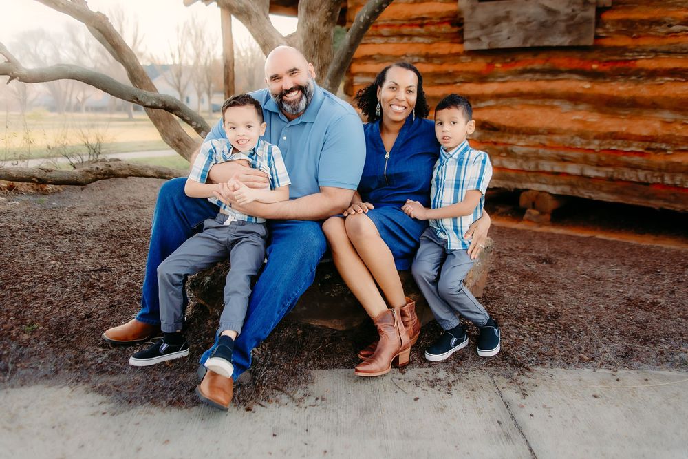 Happy family of four posing outdoors near a rustic log cabin, parents and two young boys dressed in coordinating blue outfits.