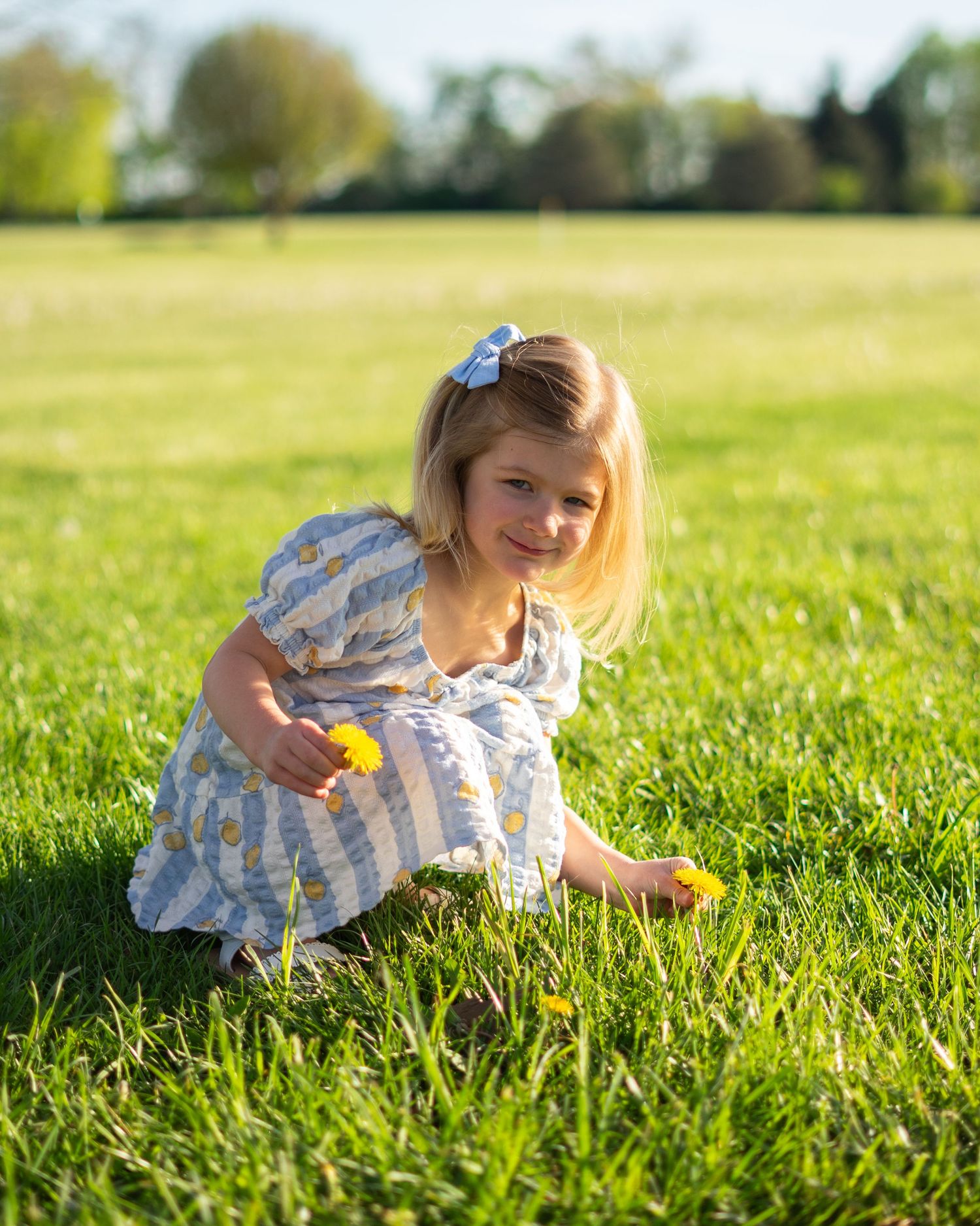 Young girl in blue floral dress picking yellow dandelions in a sunny green meadow.