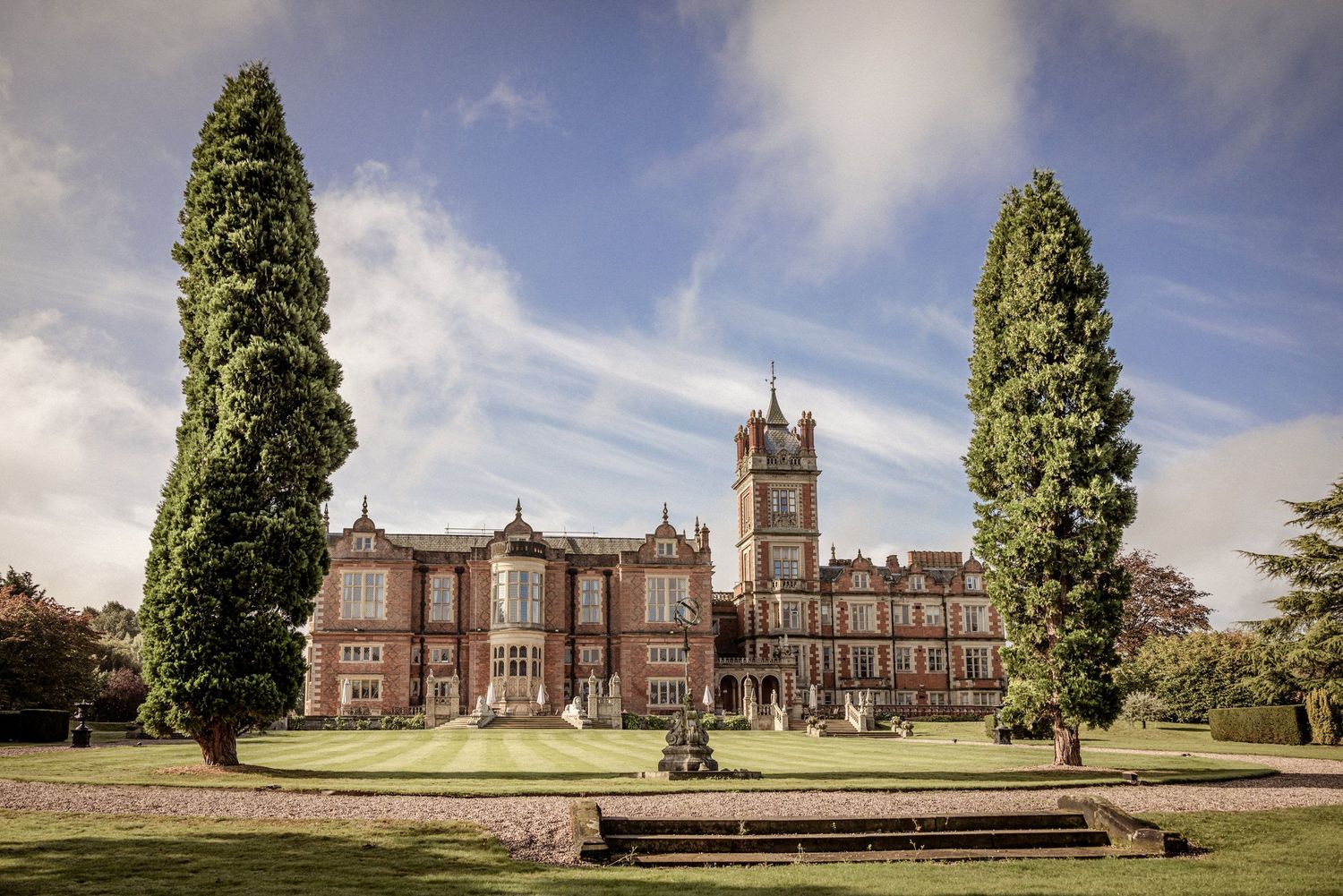 Grand Victorian red-brick manor with clock tower, flanked by tall cypress trees on manicured lawns under a blue sky.