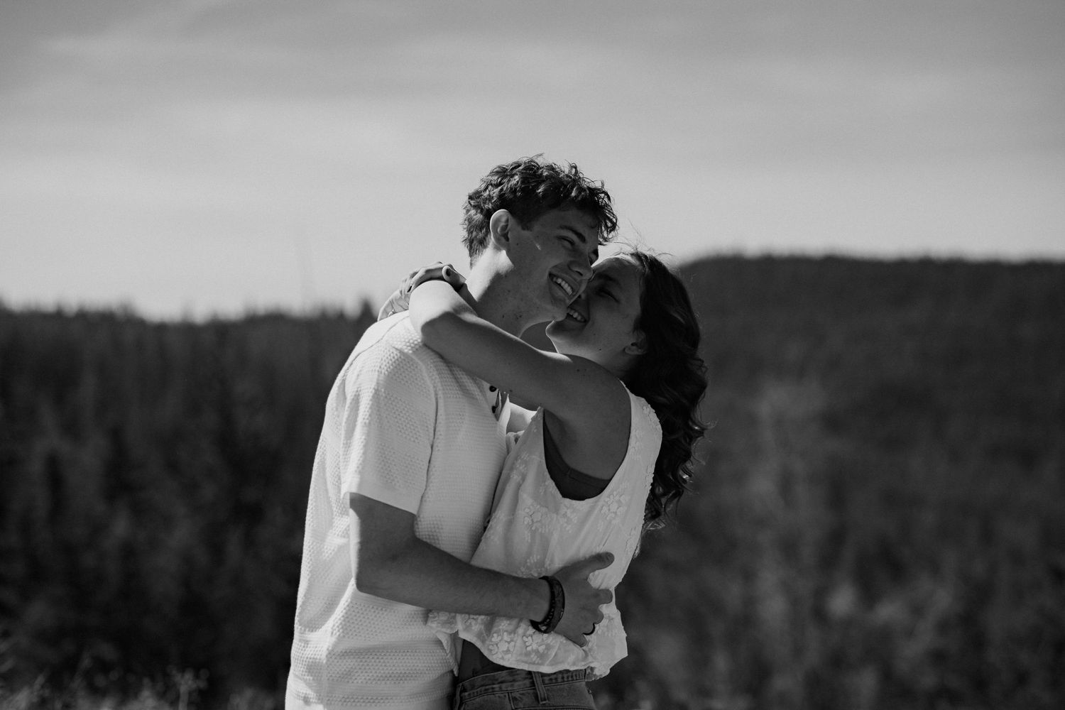 Black and white photo of a romantic couple embracing and smiling outdoors with a forest backdrop.