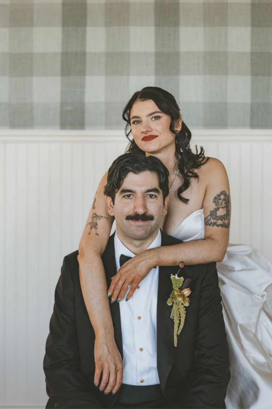 A bride and groom pose formally in wedding attire, she in a white gown with tattoos, he in a black tuxedo with boutonniere.
