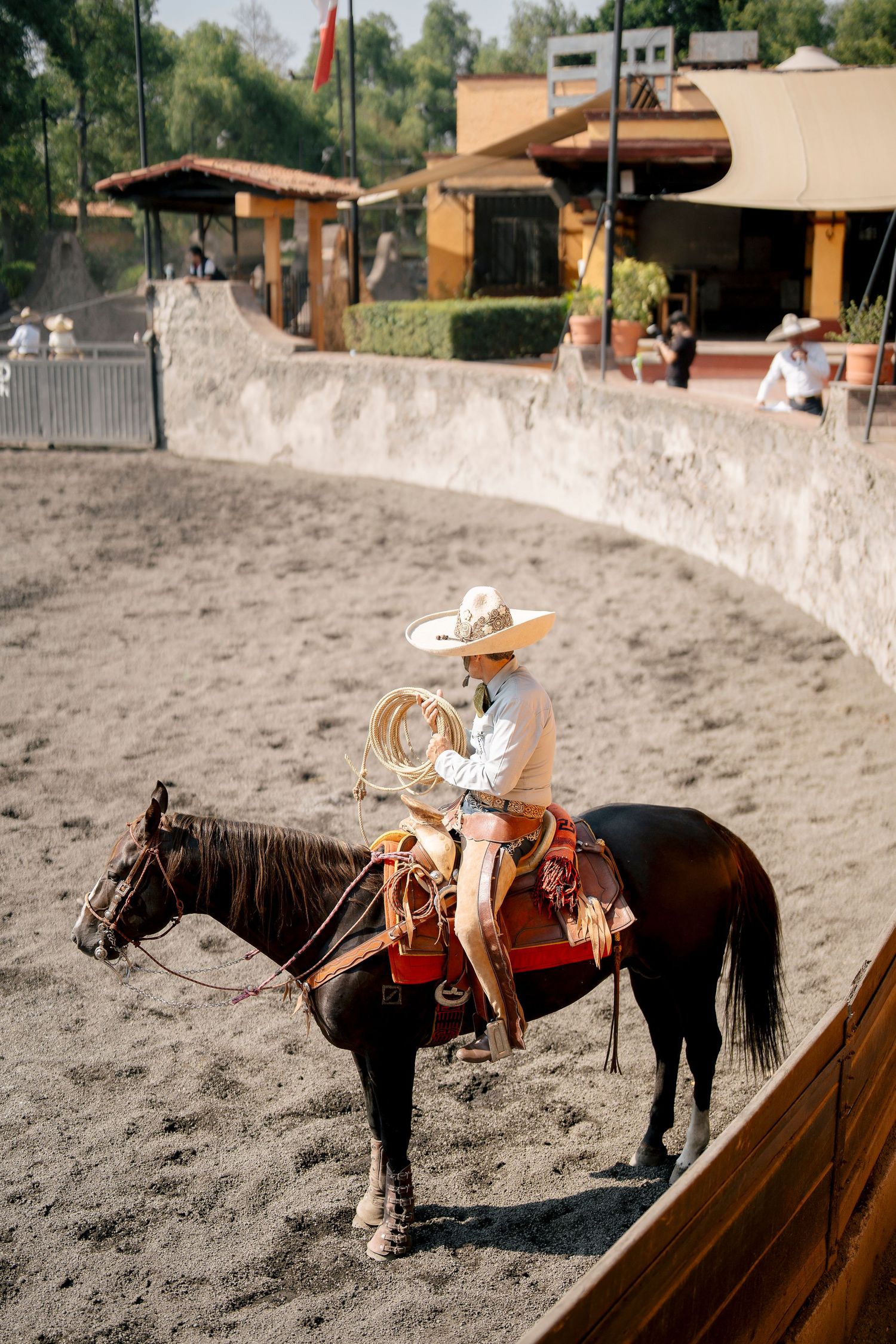 A charro in traditional attire rides a dark horse while holding a lasso in a sandy Mexican rodeo arena.