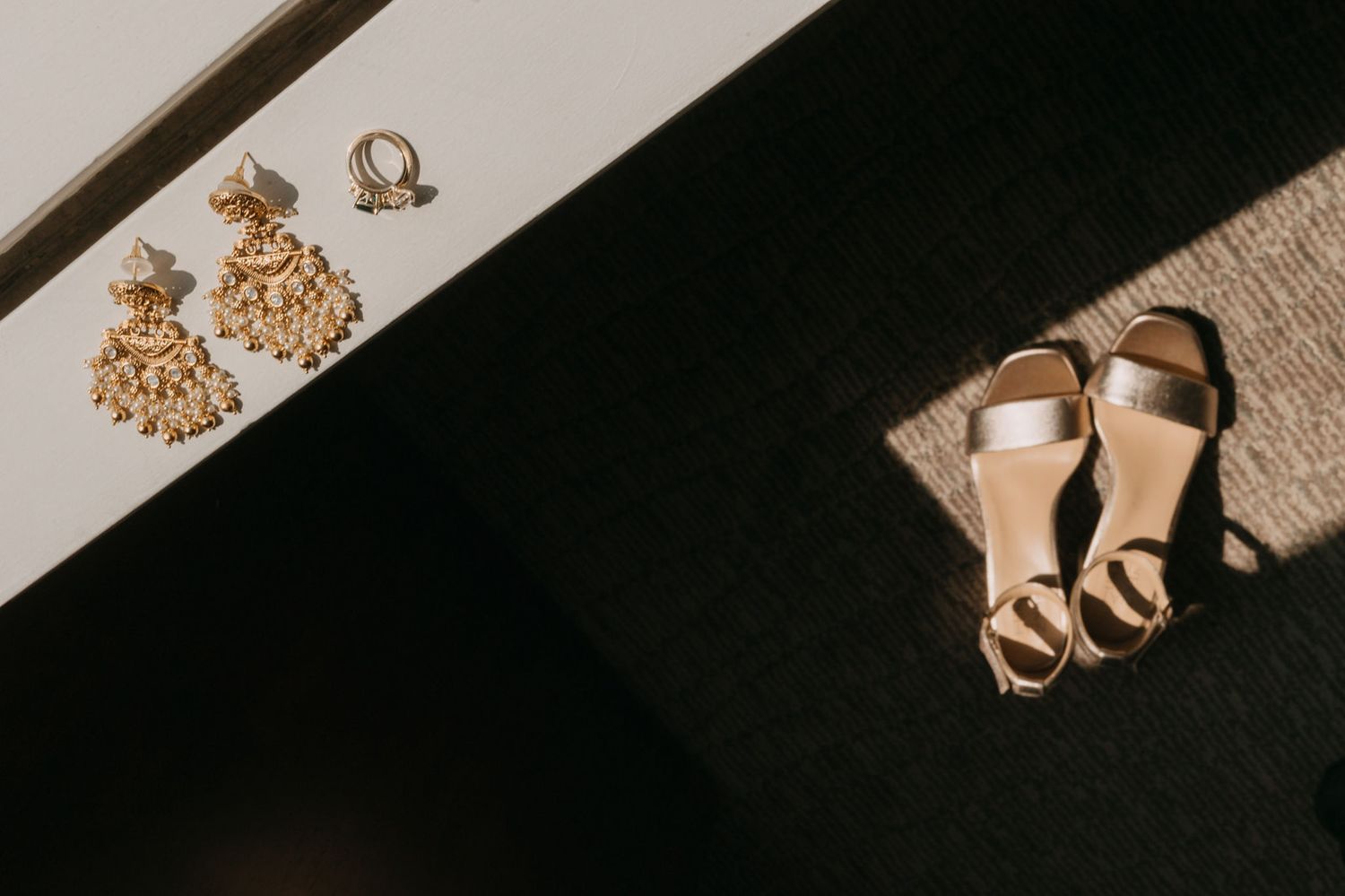 Gold chandelier earrings and strappy gold sandal heels displayed on a white surface in natural sunlight.