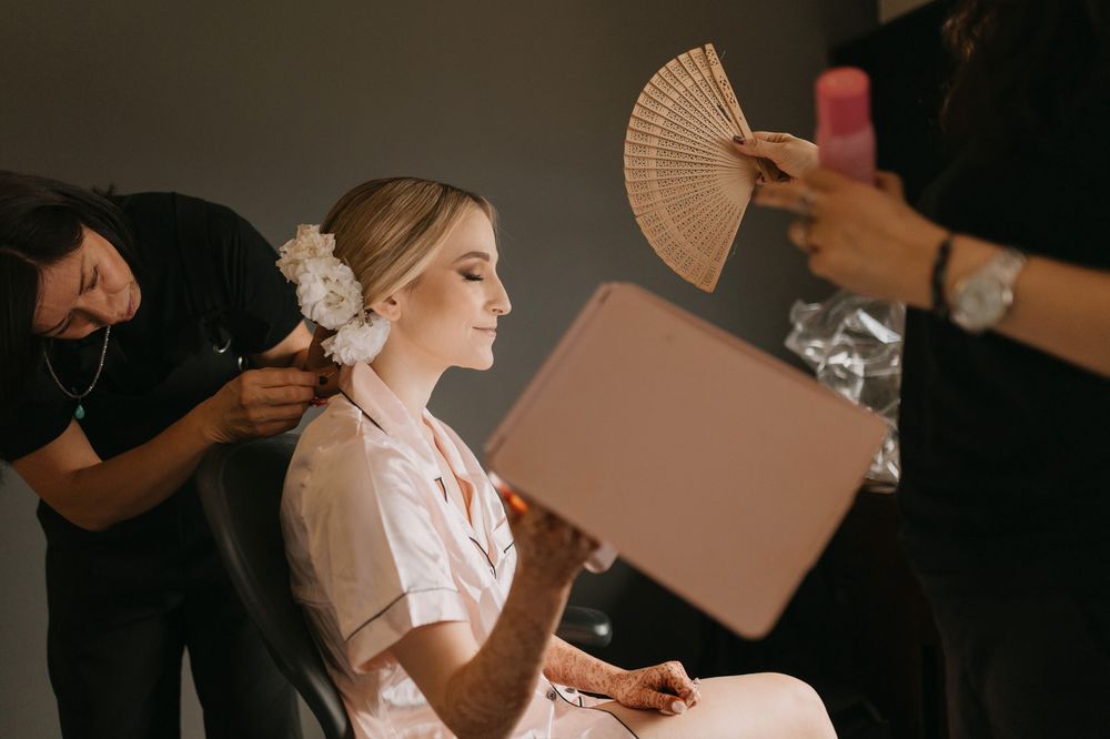 Bride getting hair and makeup done before wedding, holding a mirror while stylists work around her.