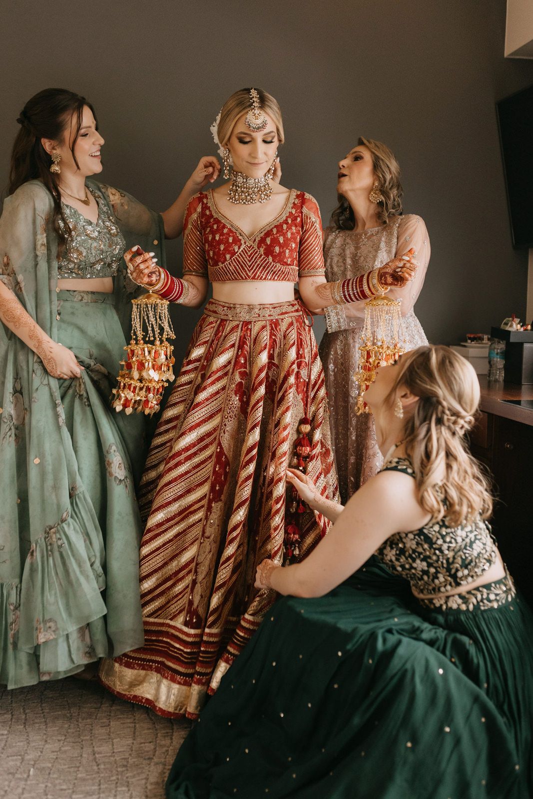 Indian bride in red lehenga surrounded by bridesmaids in green and grey outfits during wedding preparations.