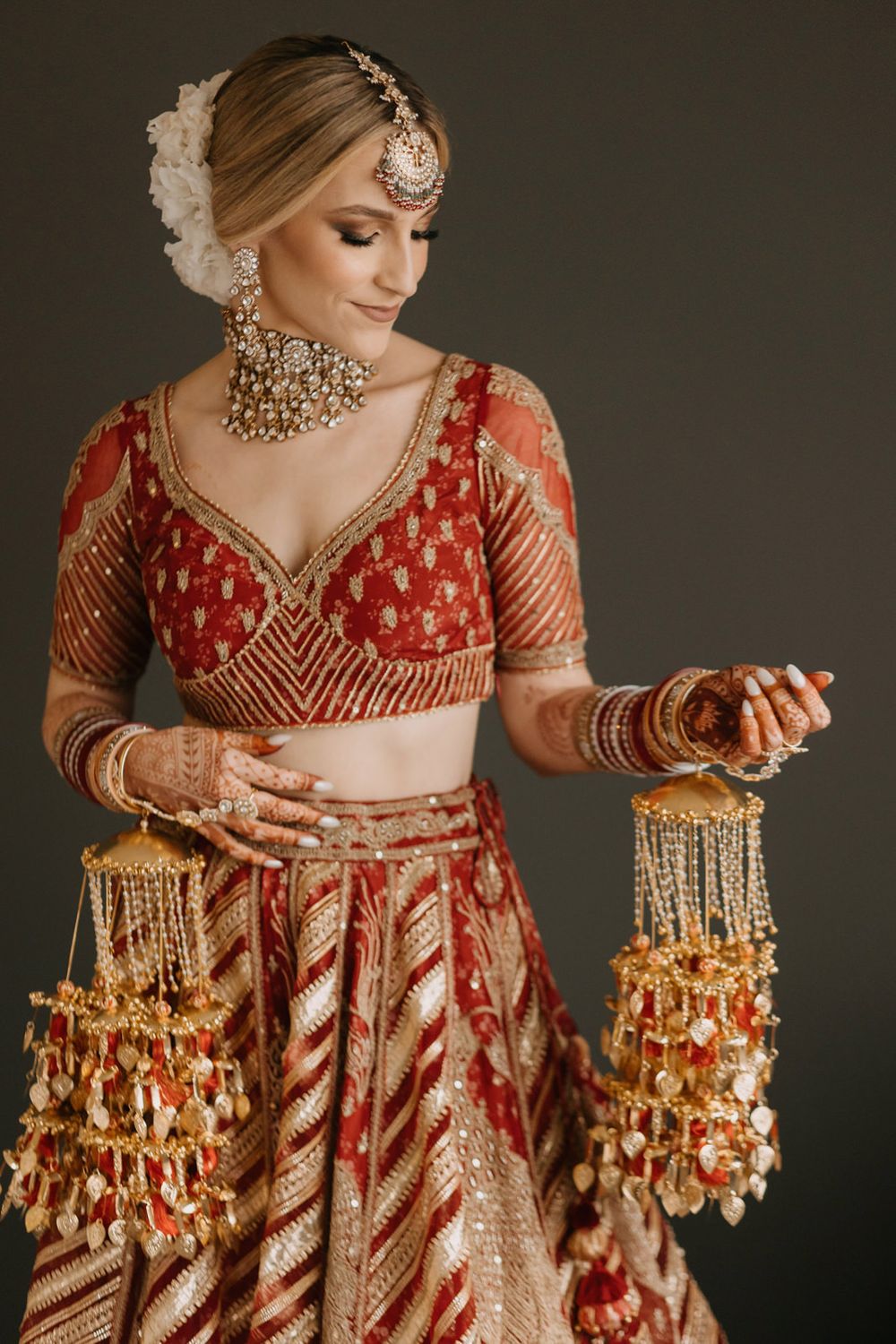 Indian bride in red lehenga holding kaleere jewelry with mehndi hands and gold kundan accessories.