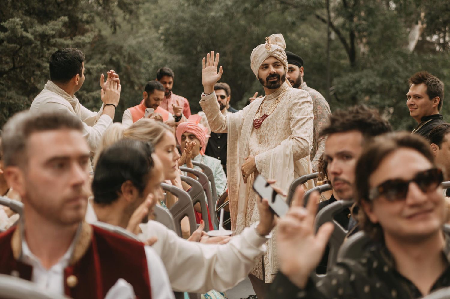 Groom in traditional Indian sherwani waves to guests at outdoor wedding ceremony surrounded by smiling attendees.