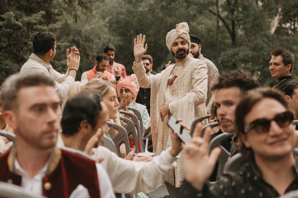 Groom in traditional Indian sherwani waves to guests at outdoor wedding ceremony surrounded by smiling attendees.