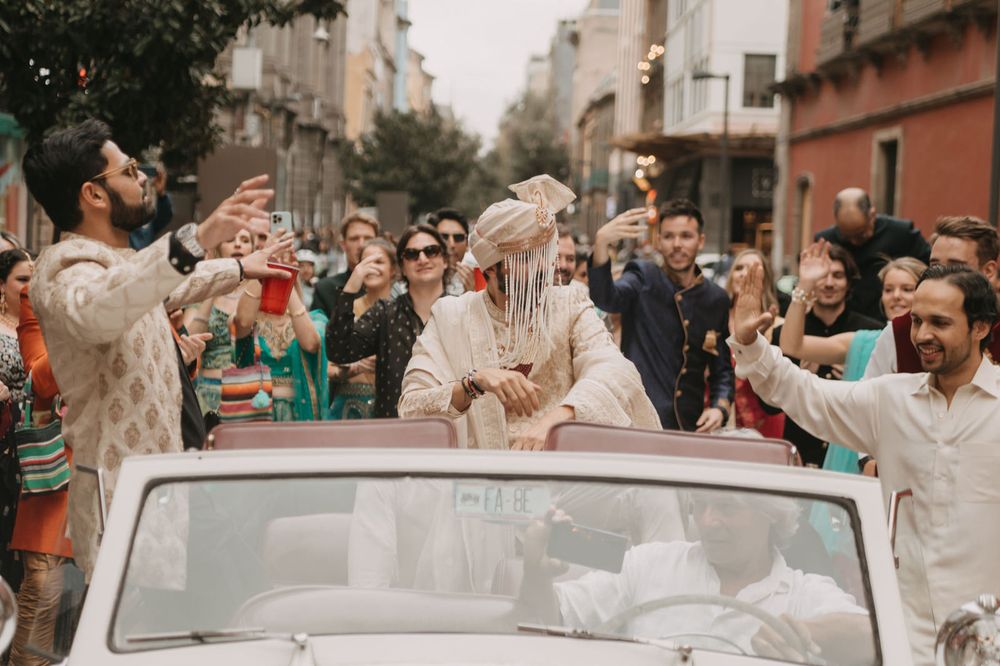 Indian groom in white sherwani and turban rides in convertible car surrounded by celebrating wedding guests on city street.