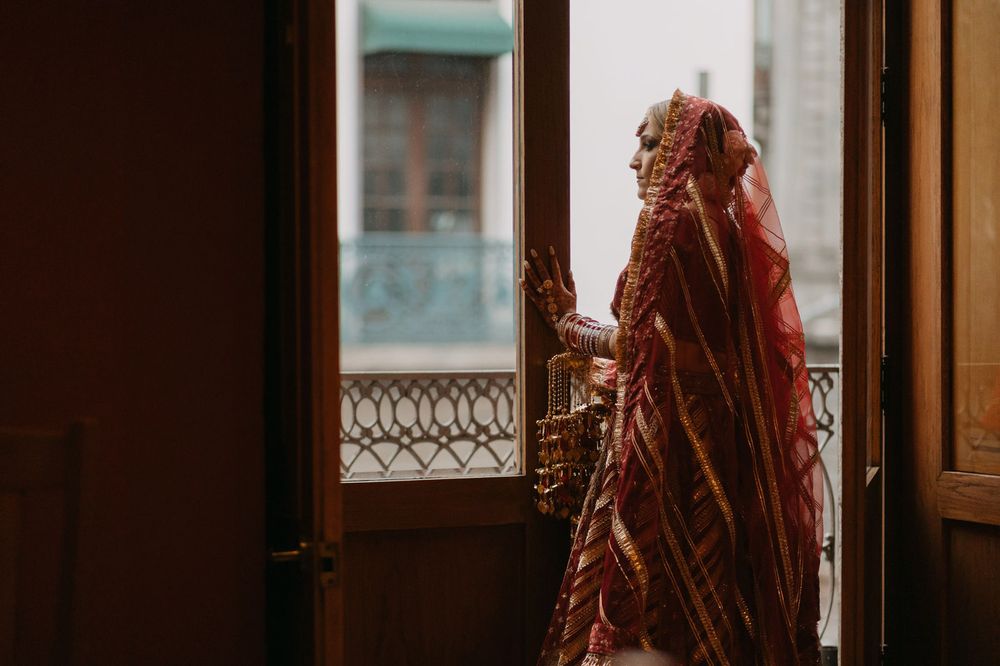 A bride in a red traditional Indian bridal outfit stands by an open balcony door in soft natural light.