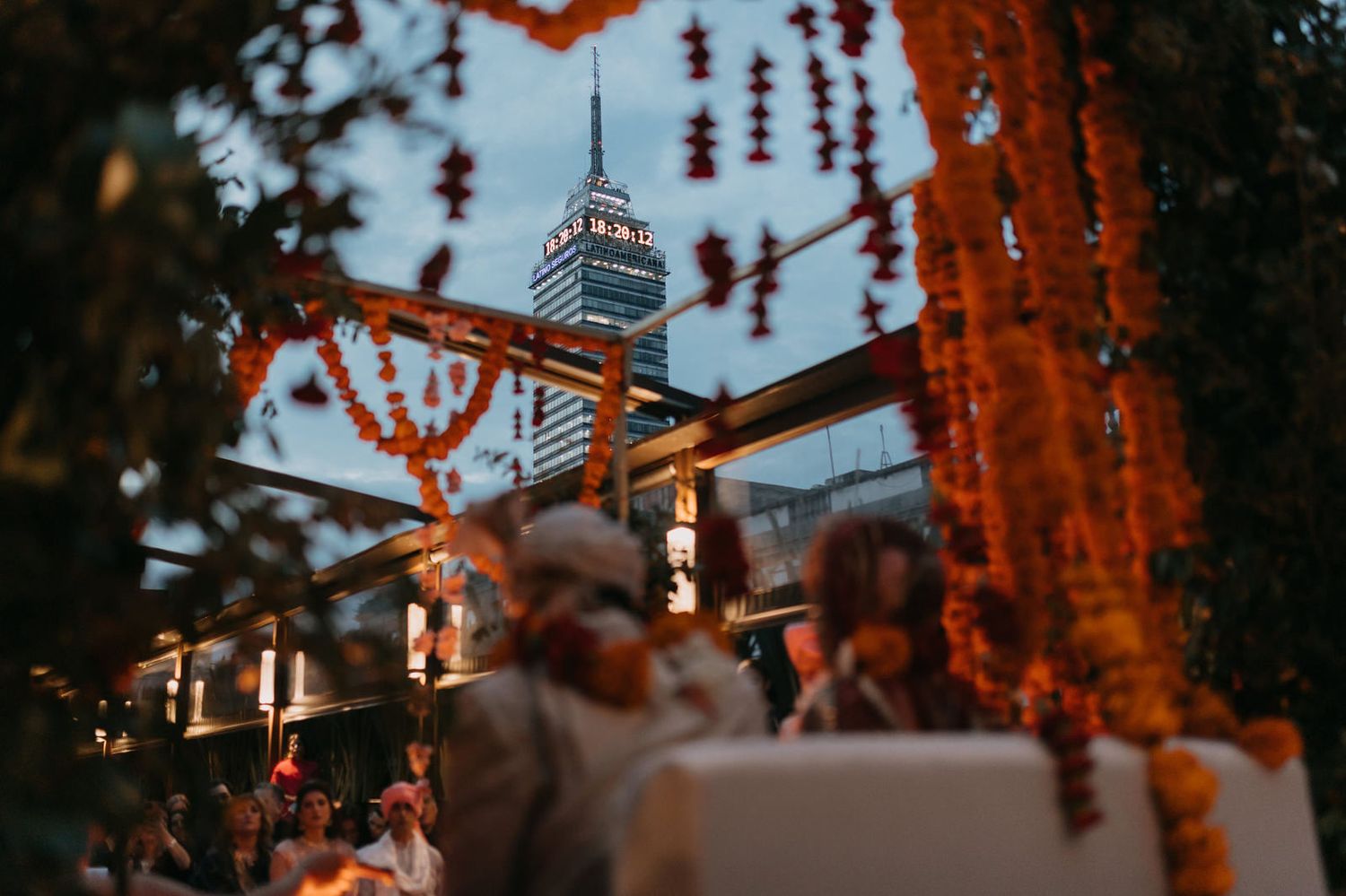 Outdoor gathering with marigold garlands framing Torre Latinoamericana skyscraper at dusk in Mexico City.