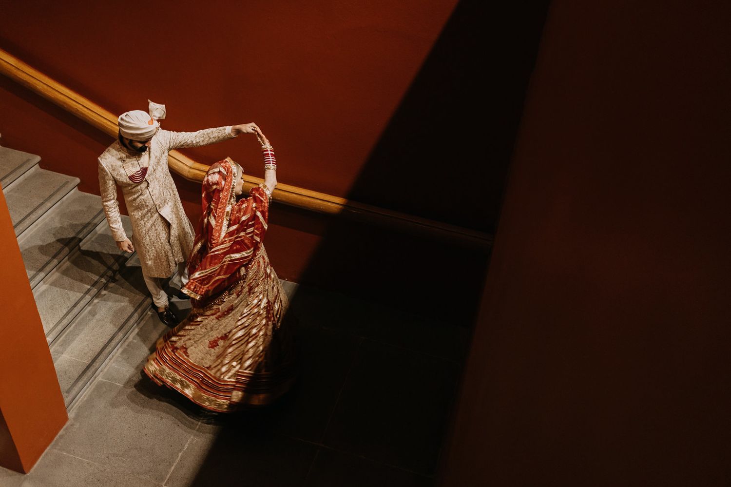 Indian bride in red saree and groom in white sherwani walking up stairs during traditional wedding ceremony.