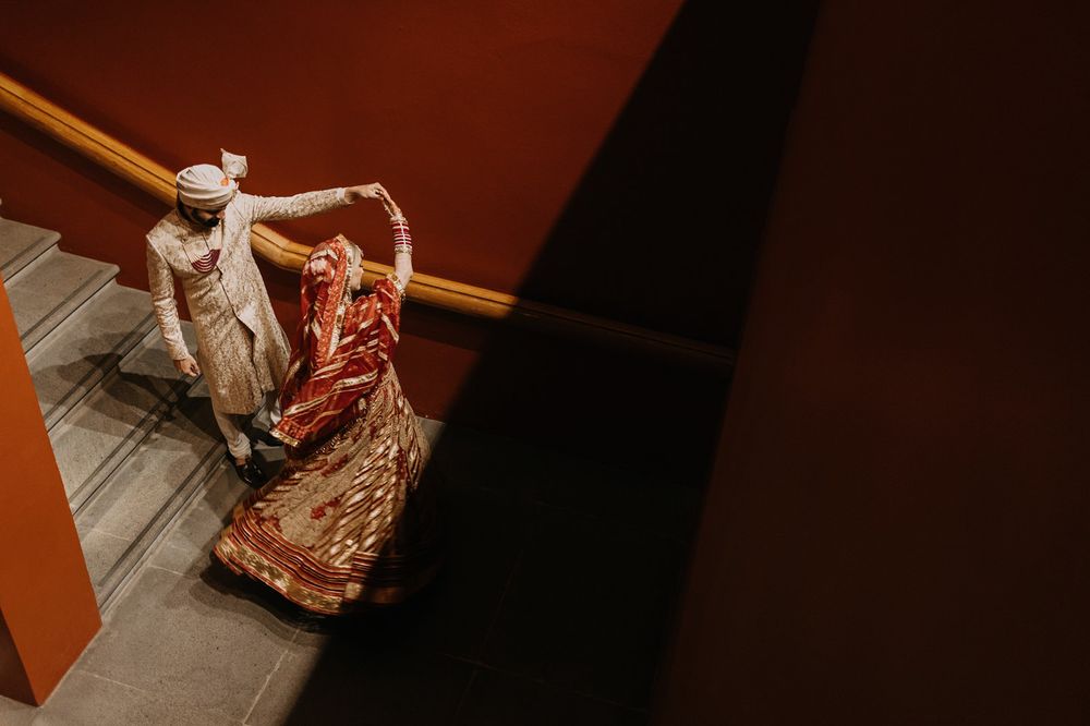 Indian bride in red saree and groom in white sherwani walking up stairs during traditional wedding ceremony.