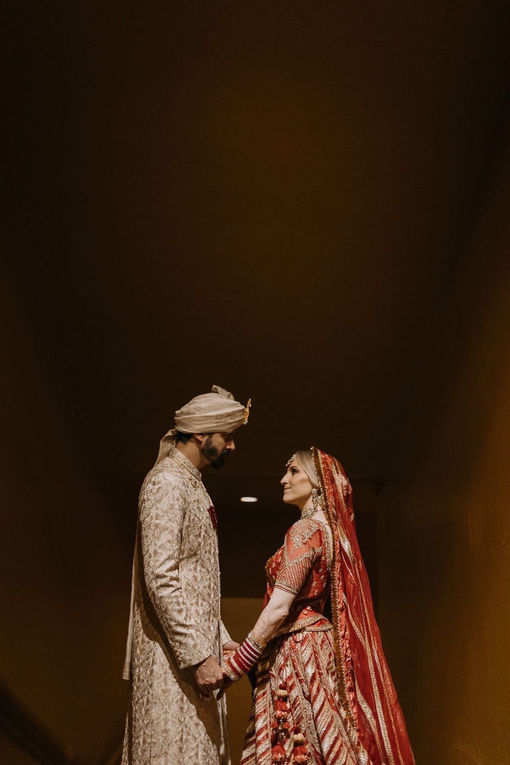 Indian bride in red lehenga and groom in cream sherwani hold hands facing each other in a romantic wedding portrait.