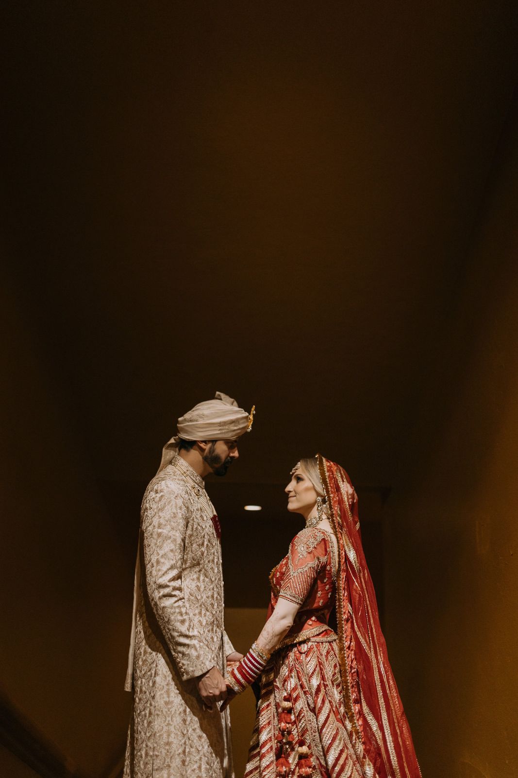 Indian bride in red lehenga and groom in cream sherwani hold hands facing each other in a romantic wedding portrait.