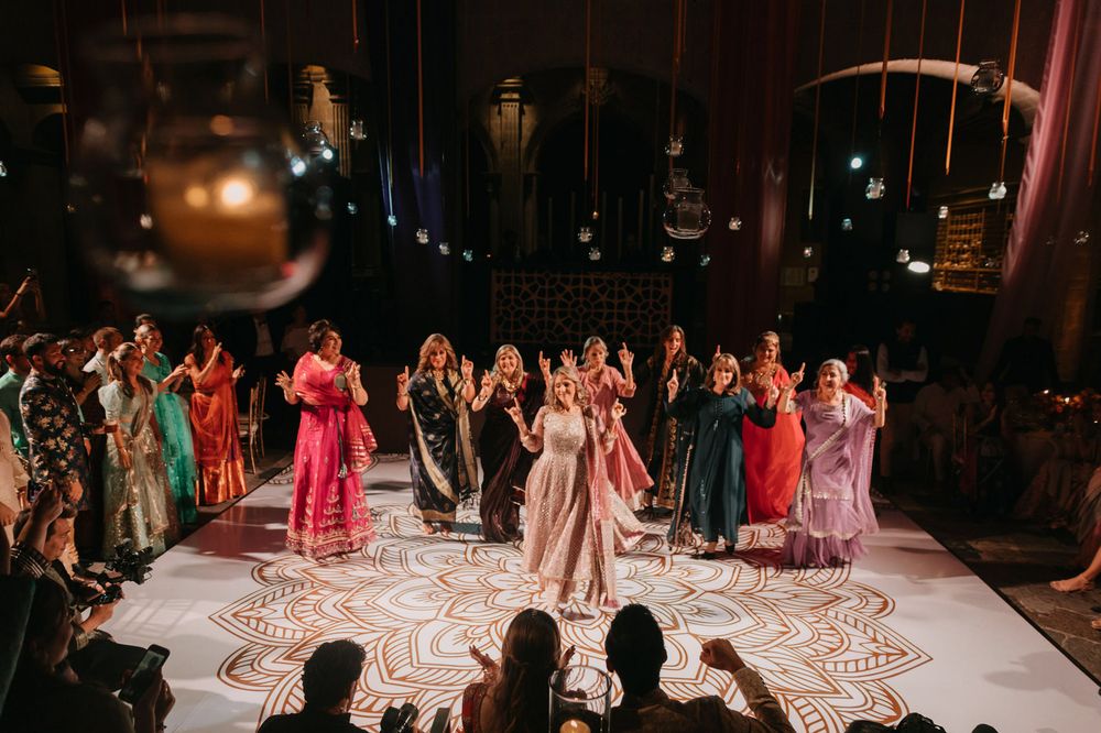 Bride in white lehenga dancing with guests in colorful ethnic wear on ornate mandala dance floor at Indian wedding.