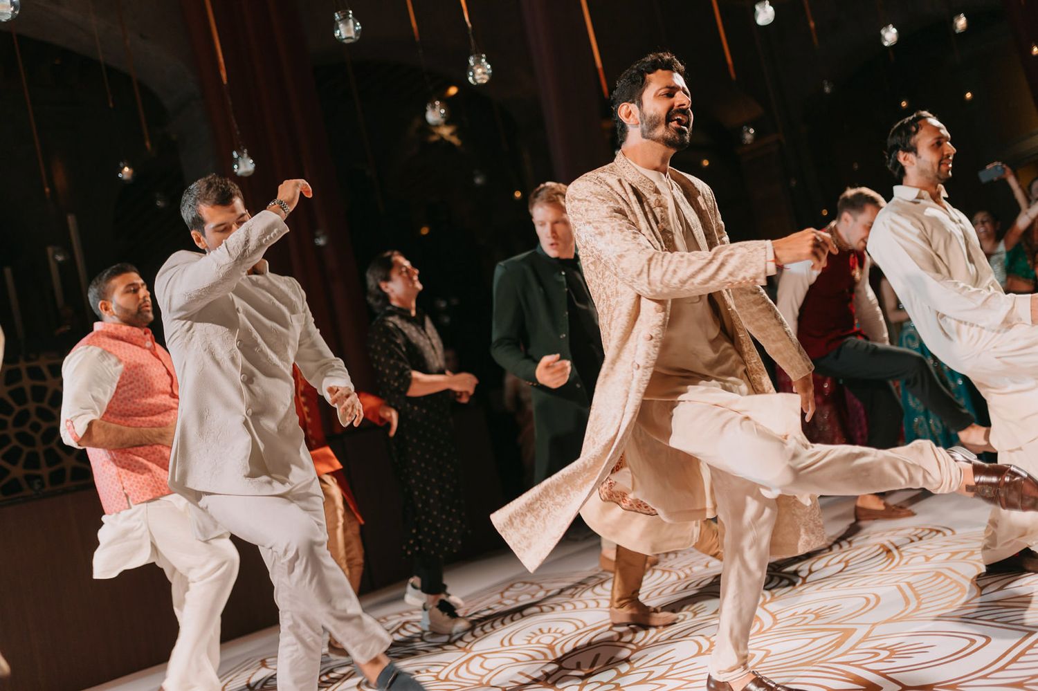 Men in traditional South Asian attire perform energetic dance moves at a festive wedding celebration.