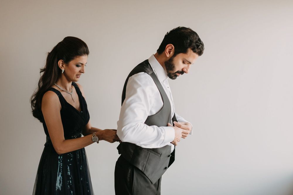 A woman in a black dress helps a bearded man in a gray vest and white shirt button his cufflinks before a formal event.