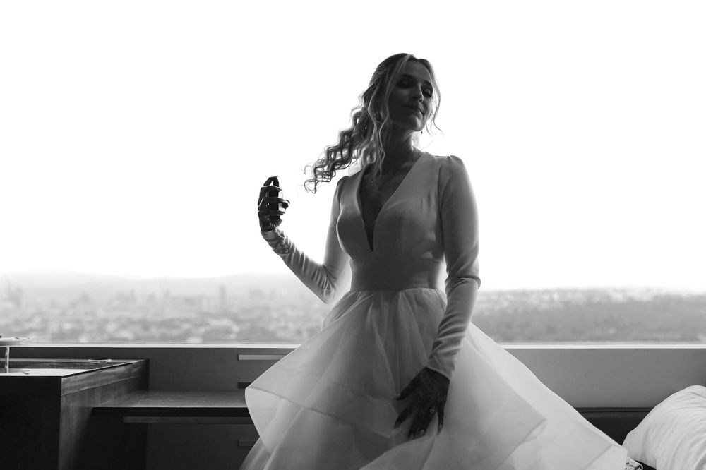 Black and white photo of a bride in a white tulle gown with long sleeves posing by a window with city views.