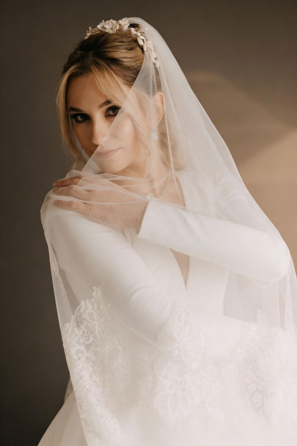 Elegant bride in a white lace gown and floral headpiece, draped in a sheer veil, posing gracefully indoors.
