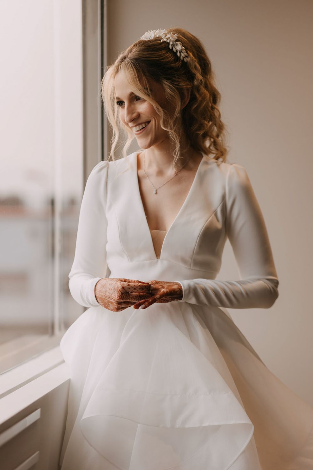 Smiling bride in a white long-sleeve deep V-neck wedding gown with hair accessory stands by a window.