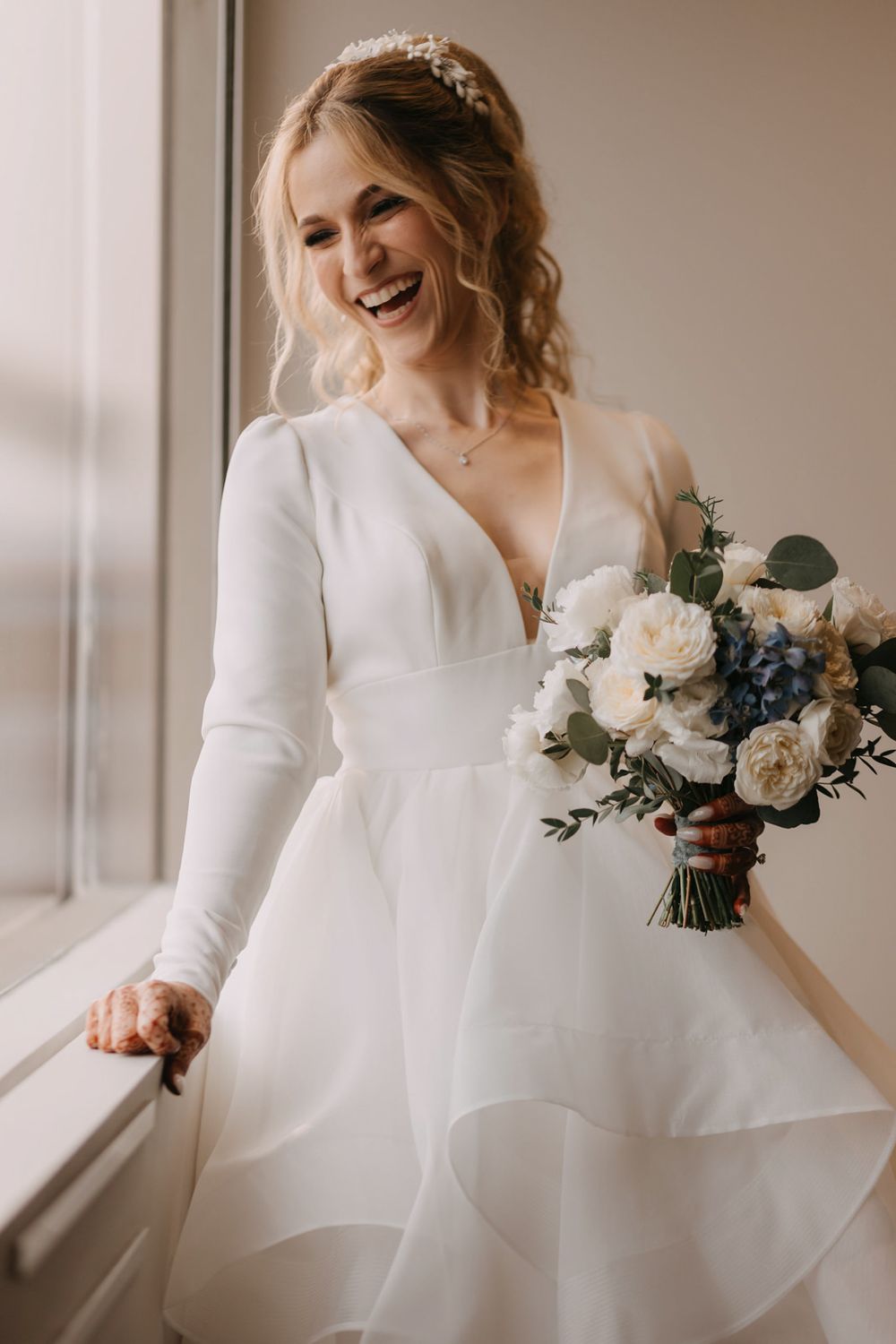 Smiling bride in long-sleeve white wedding gown holds bouquet of white and blue flowers near a bright window.