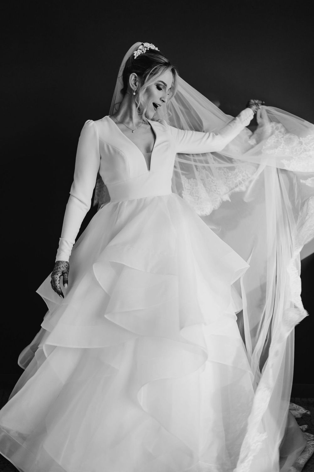 Black and white photo of a bride in a layered tulle gown with long sleeves, holding her veil dramatically.