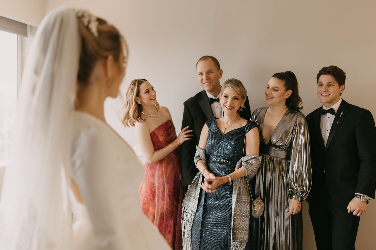 Bride in white gown and veil shares emotional first look moment with smiling family members in formal attire.