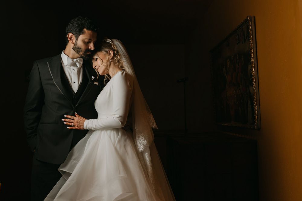 Elegant bride and groom share an intimate moment in dramatic lighting, bride in long-sleeve white gown with veil.