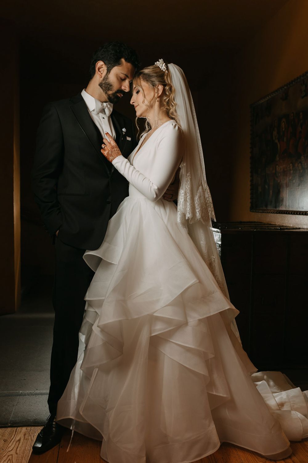 Bride in layered white gown and veil stands close with groom in black tuxedo in a dimly lit elegant room.