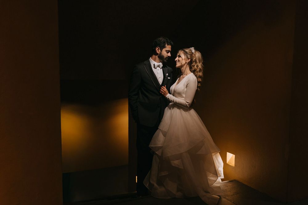 Romantic wedding couple in elegant attire share an intimate moment in a warmly lit corridor.