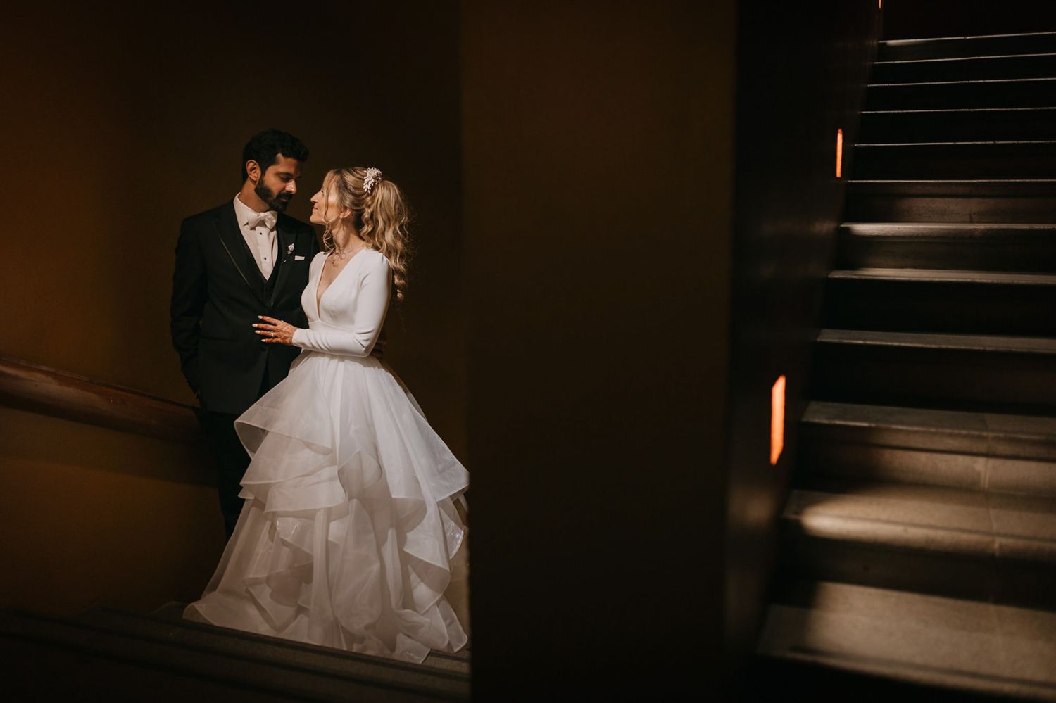 Bride and groom share romantic moment in dimly lit stairwell, bride in elegant white gown with long sleeves.