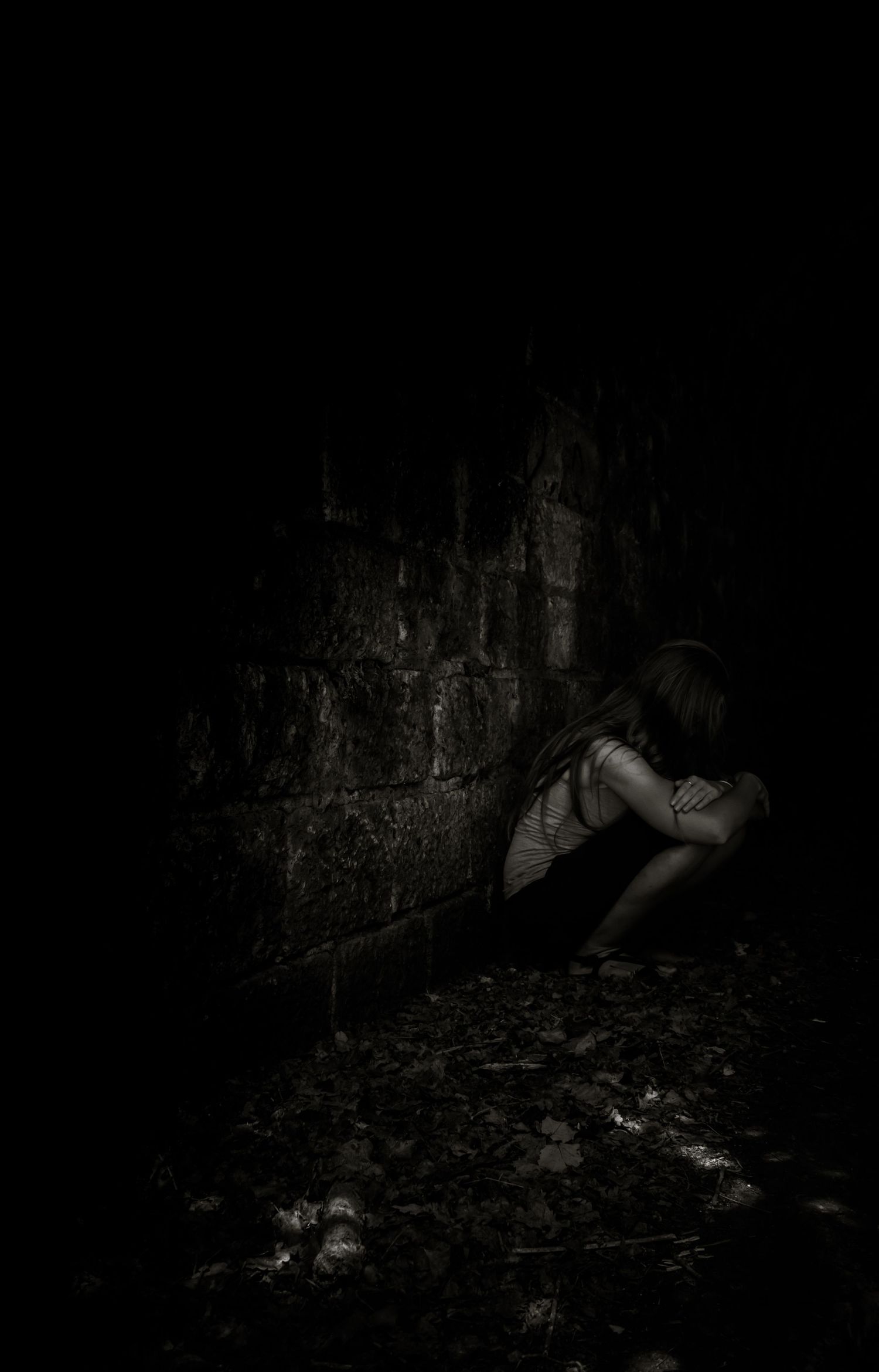 Une jeune femme est accroupie seule dans un coin sombre et abandonné, adossée à un mur de pierre, sur une photo en noir et blanc.