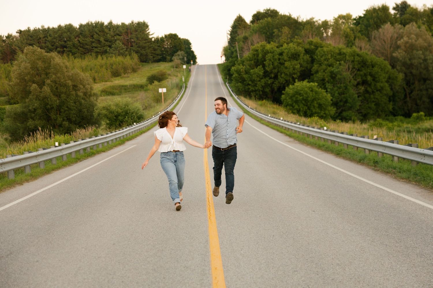 Couple holding hands and running together on an empty road surrounded by lush green trees.