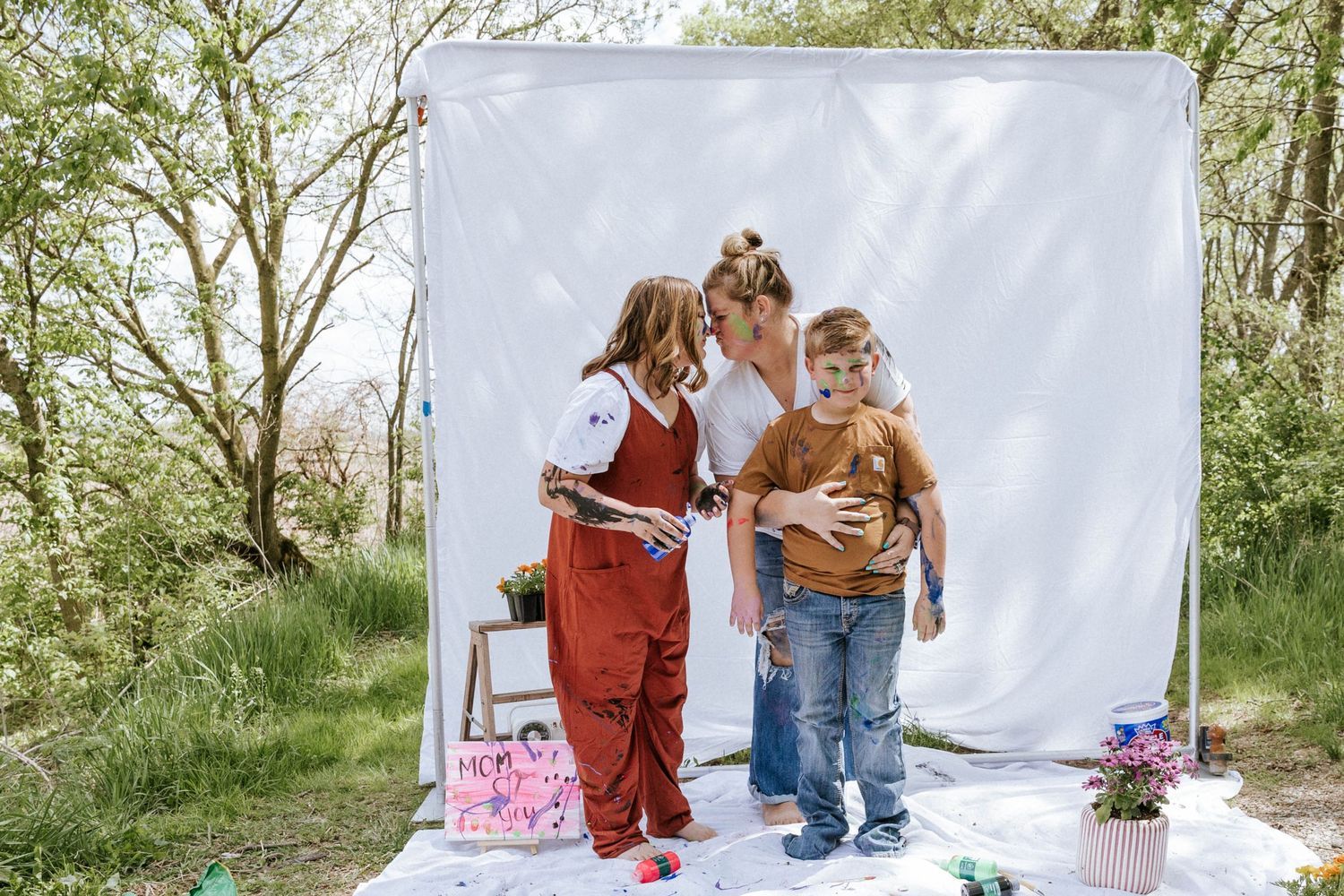 A mother and two young children pose in front of a white backdrop outdoors among trees.