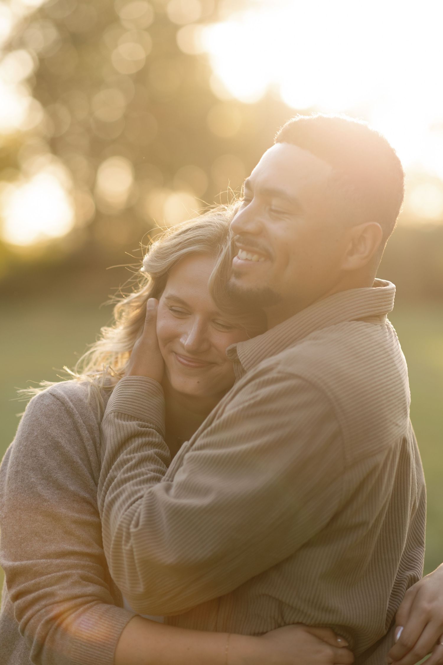 Couple embracing warmly at golden hour sunset in a field, smiling contentedly in matching earth-tone sweaters.