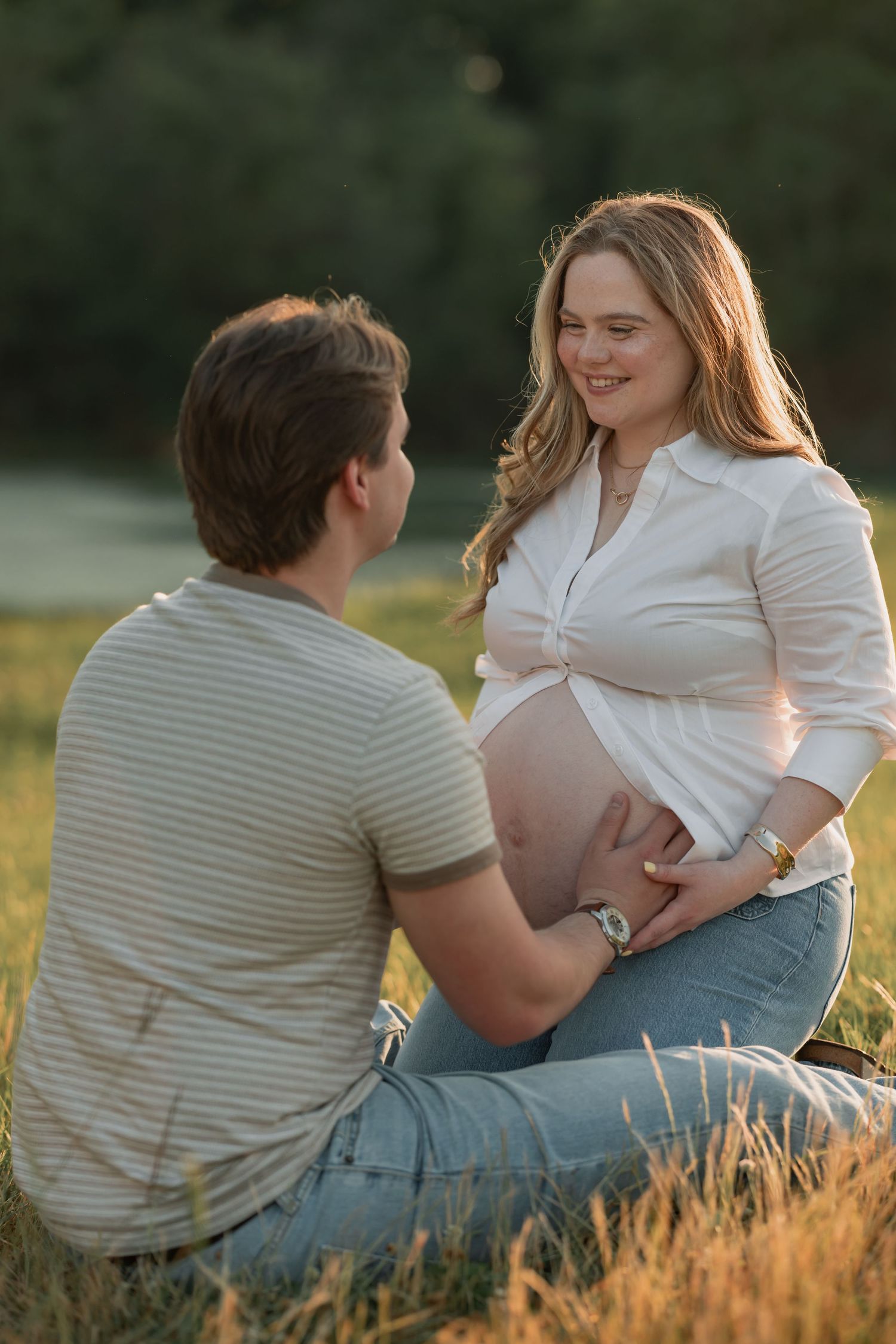 Pregnant woman in white shirt sits with partner in golden field, both touching her baby bump at sunset.
