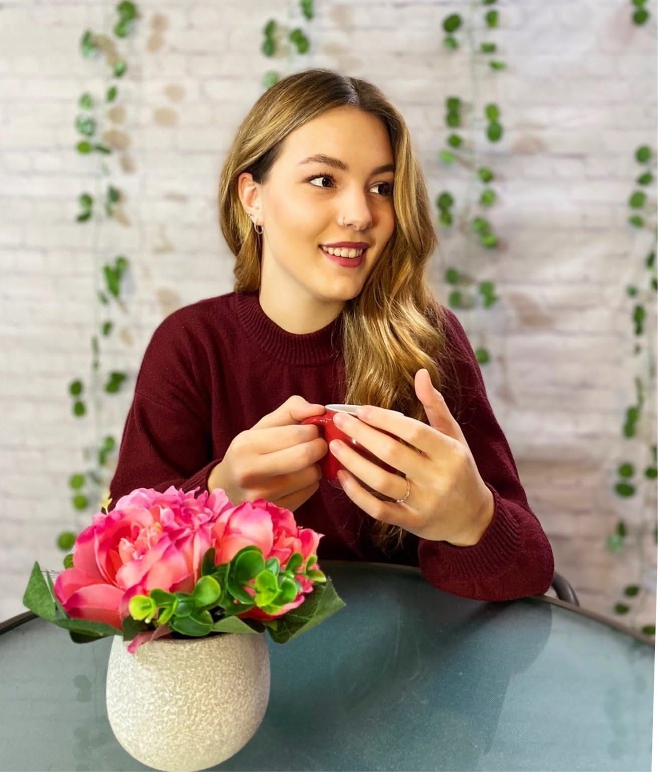 Smiling young woman in burgundy sweater sits at glass table with pink roses in white vase indoors.