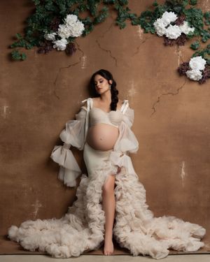 Elegant maternity portrait with ruffled white gown against brown wall with floral accents.